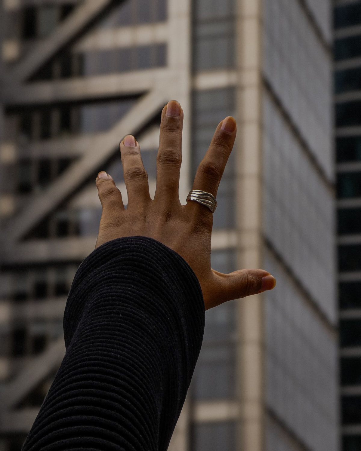 A hand reaching up towards City of London towers, wearing the Blurr ring in recycled sterling silver